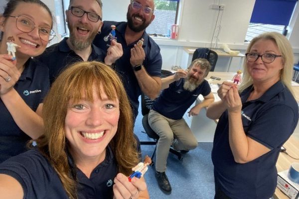 Six Proactive colleagues taking a group selfie in the office, each holding up a small toy figure