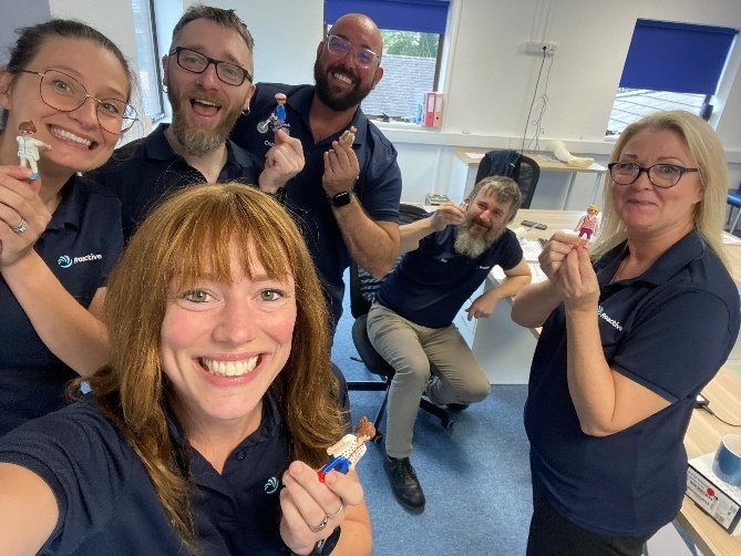 Six Proactive colleagues taking a group selfie in the office, each holding up a small toy figure