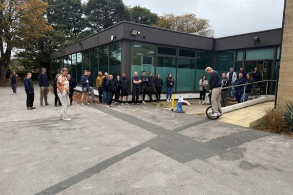 A colleague attempting the unicycle challenge while the team watches outside the building