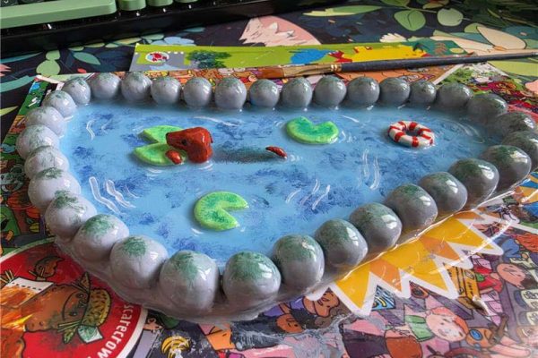 Amy's finished pond ring dish, painted blue with green lily pads, a red creature swimming and a red and white rubber ring