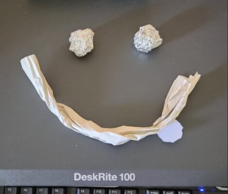 Smiley face on a desk mat, made from two balls of foil for eyes and a twisted strip of paper for a mouth