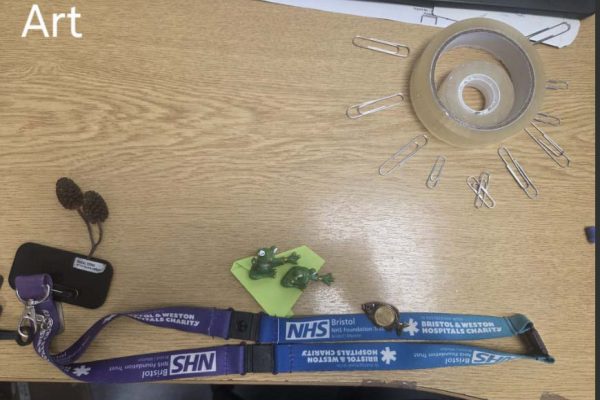 Labelled "Art": a desk scene with a sun made from a roll of tape and paperclips, two toy frogs on a sticky note, and NHS lanyards
